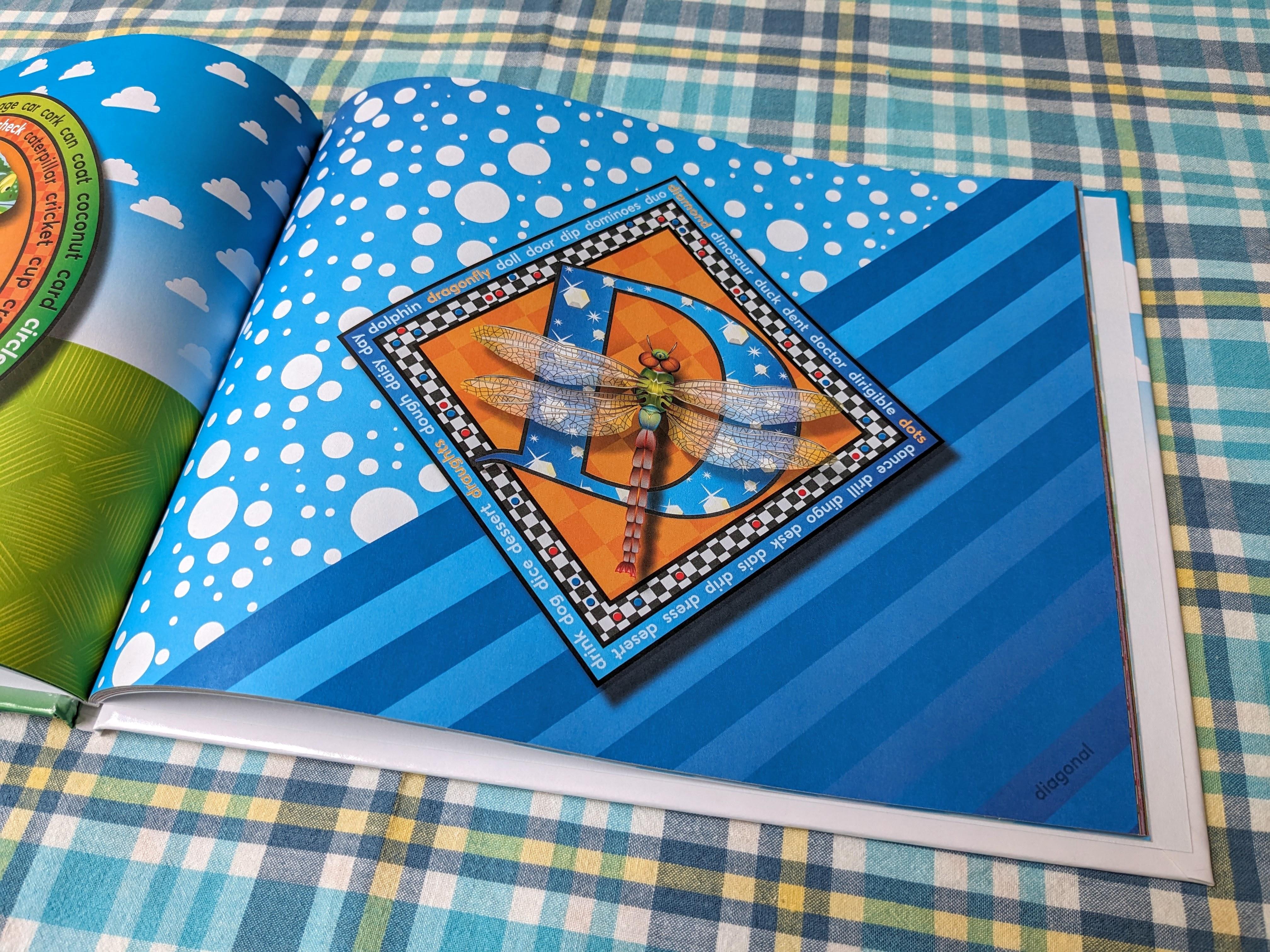 Photograph of the open alphabet book. The right page features an orange letter D with a large, detailed, multi-coloured dragonfly resting on it. The background consists of blue and white polka dots and diagonal blue stripes.
