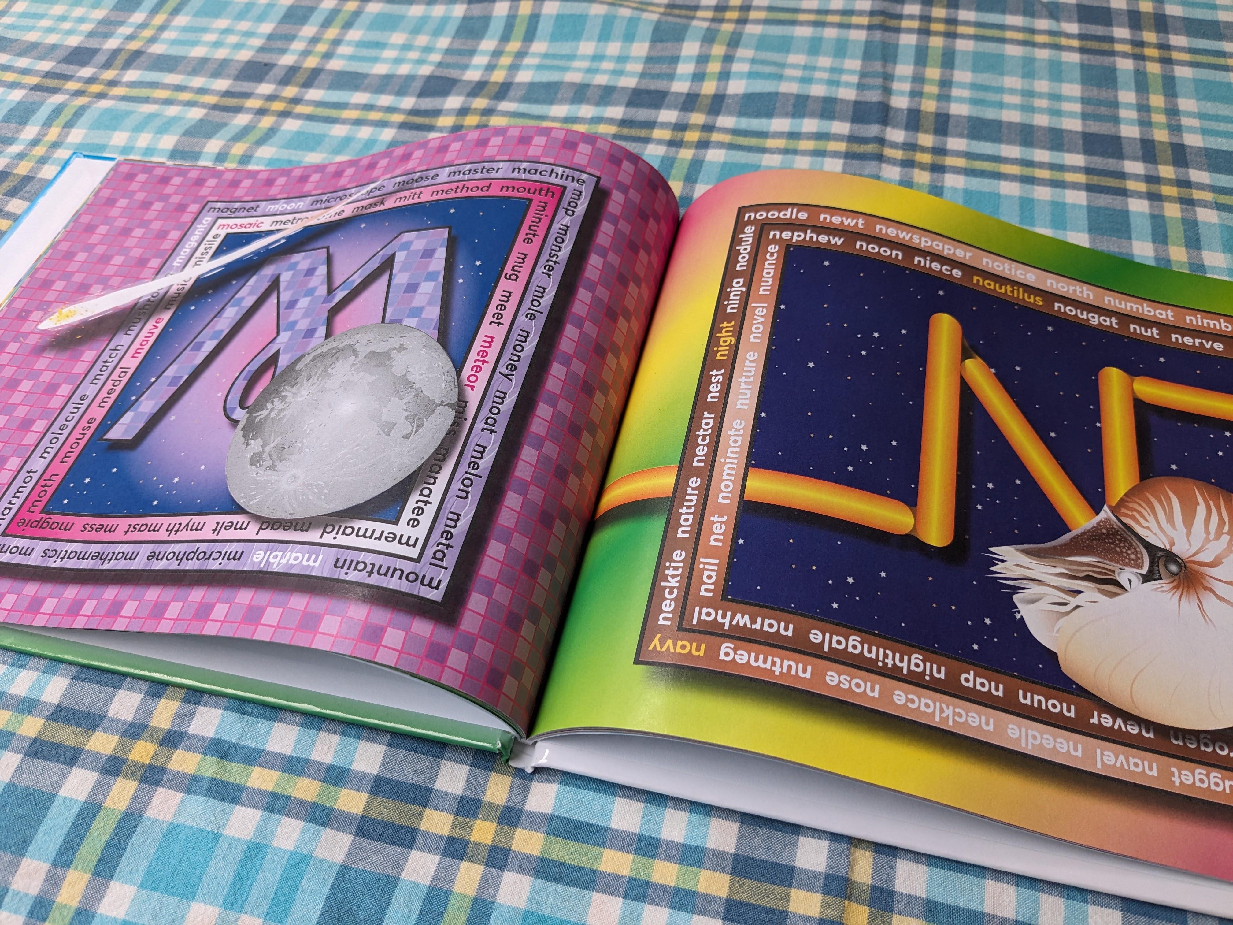 Photograph of the open alphabet book. The left page shows a purple patterned letter M with an illustration of the full moon. The right page shows a yellow letter N made of intersecting tubes alongside a large nautilus shell.