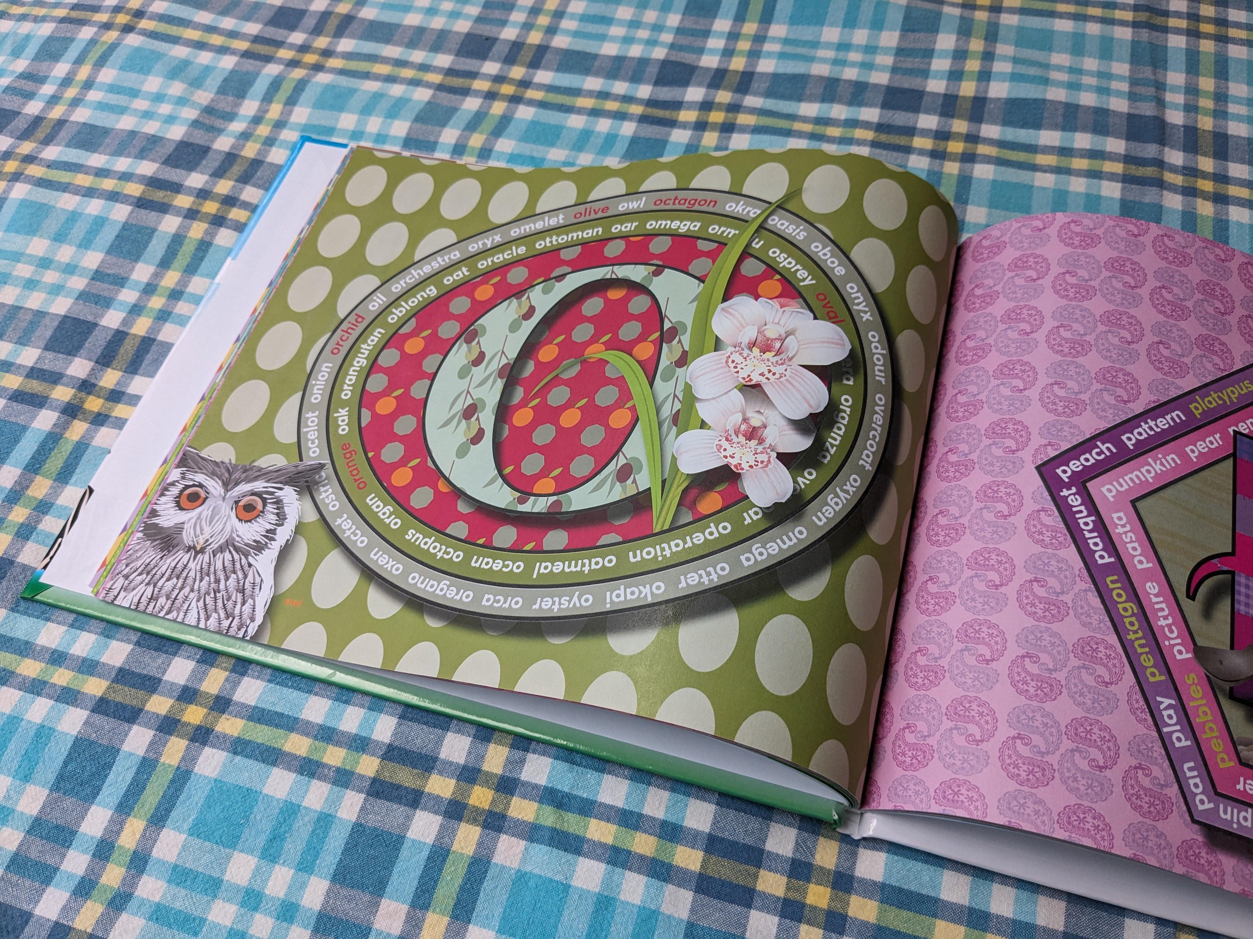 Photograph of the open alphabet book. The left page features a large red letter O with a polka dot pattern, surrounded by white orchid flowers and green leaves. An illustration of an owl peers in from the bottom left corner.