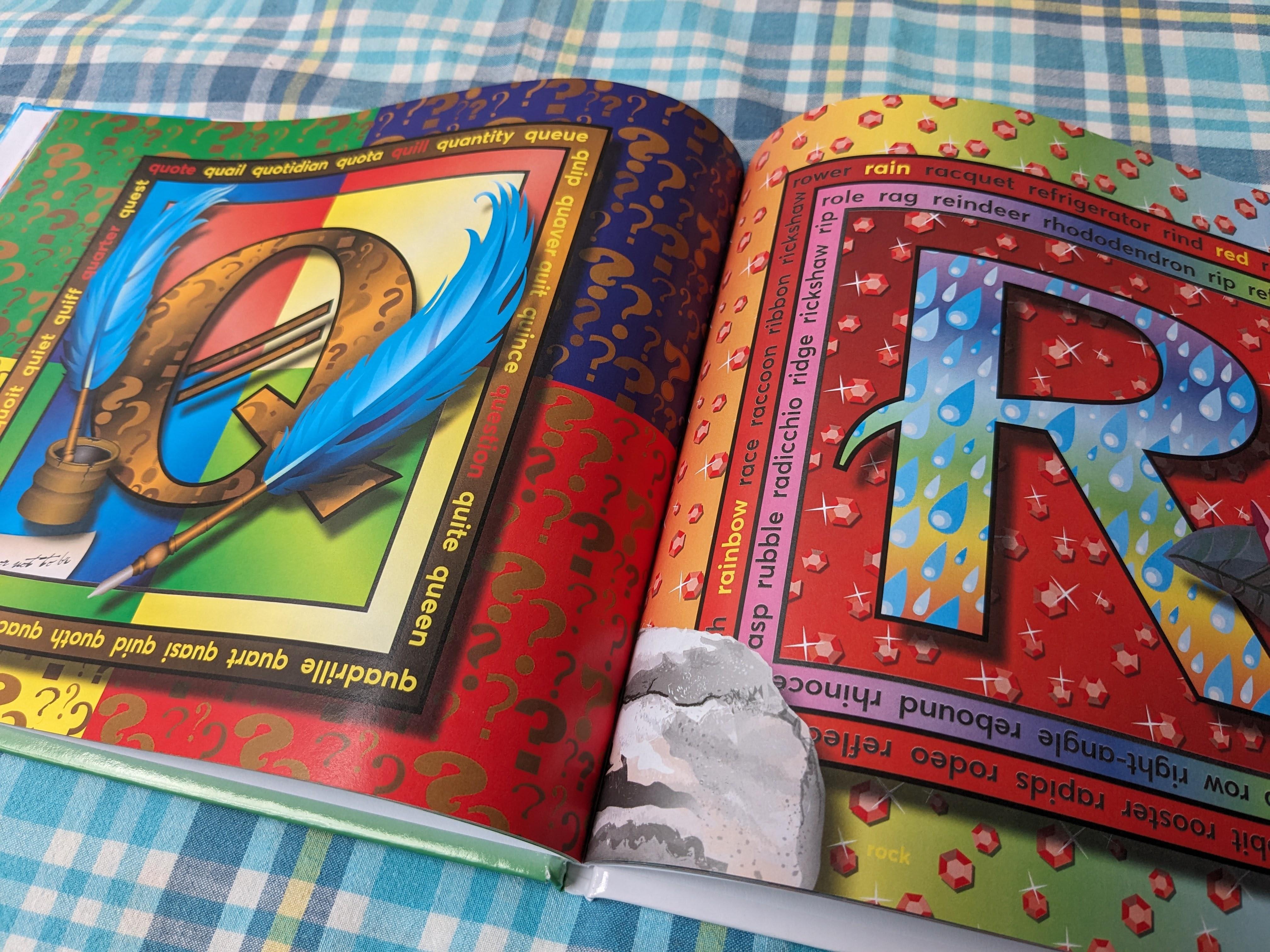 Photograph of the open alphabet book. The left page shows a gold letter Q crossed by two blue feather quills. The right page shows a blue letter R patterned with raindrops next to a red border and background patterned with red jewels.